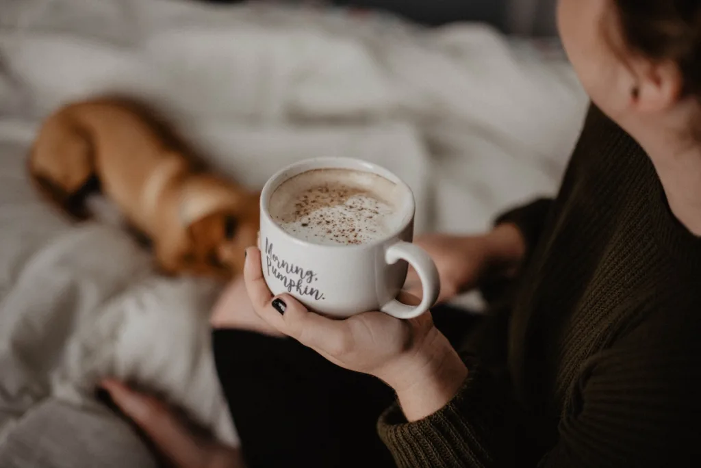A woman with a cup of coffee sitting in bed with her dog