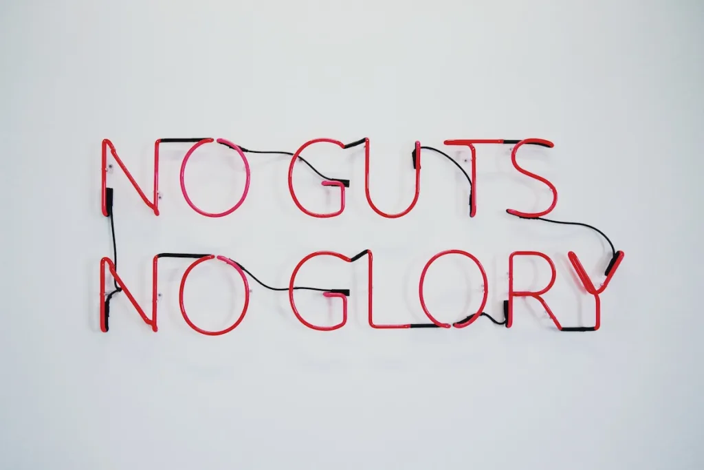 A red neon sign on a white background. The sign says "no guts, no glory".