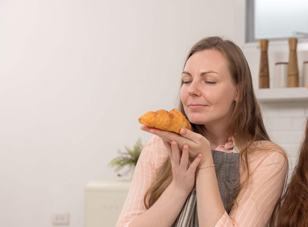 A woman who is practicing intuitive eating by being mindful. She is holding a plate with a croissant roll with her eyes closed