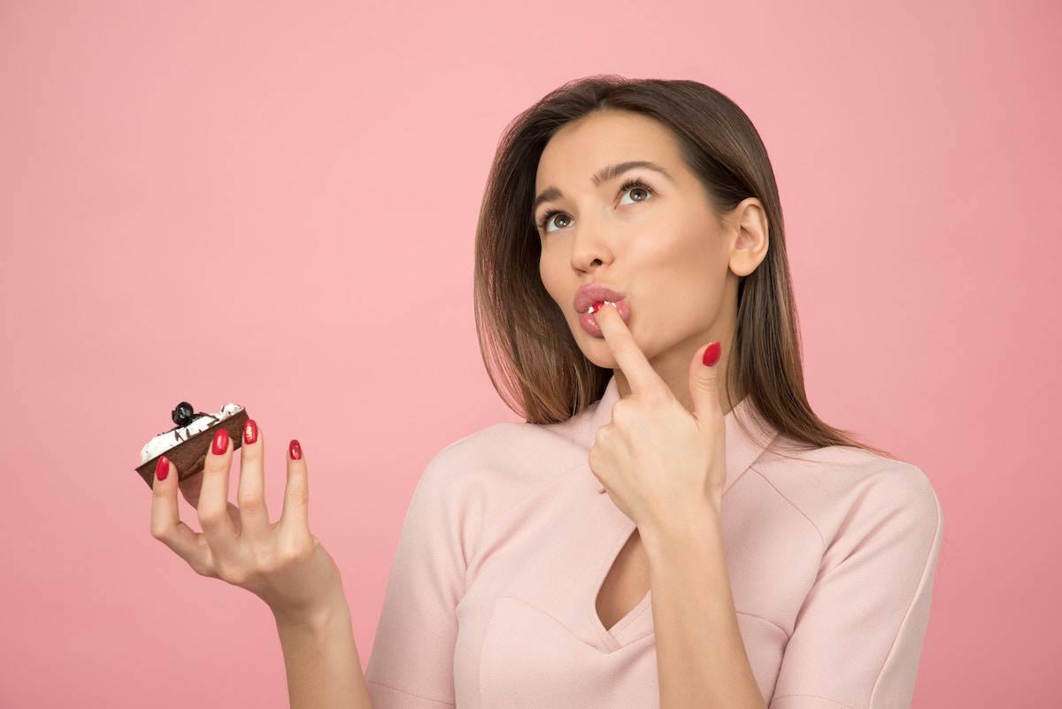 A woman with a pink background eating a cupcake and licking her finger.