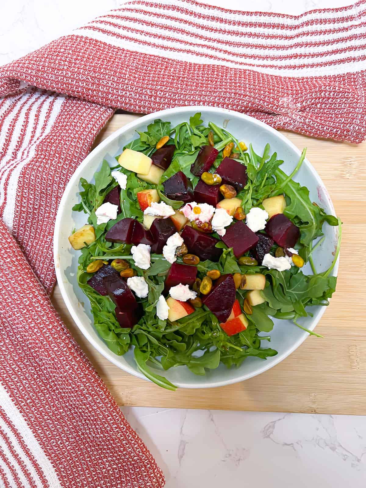 Beet and arugula salad with goat cheese in a white bowl with a red and white cloth and a wooden cutting board