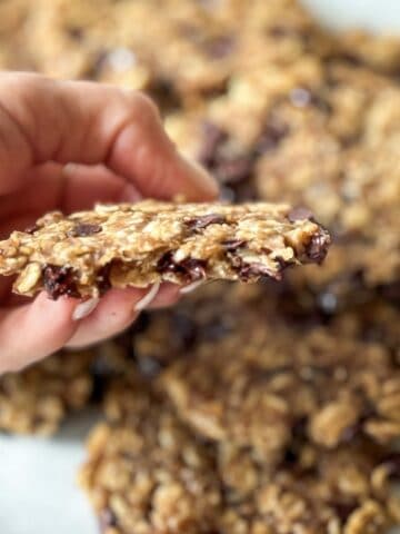 A human hand holding up a 4 ingredient banana oatmeal cookie with a plate of cookies in the background.