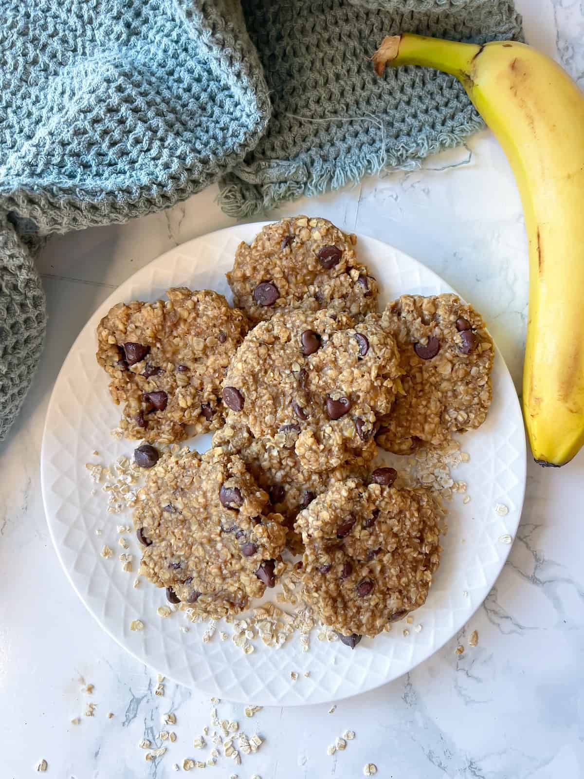 4 ingredients banana oatmeal cookies on a plate with oats and banana in the background on a white marble table
