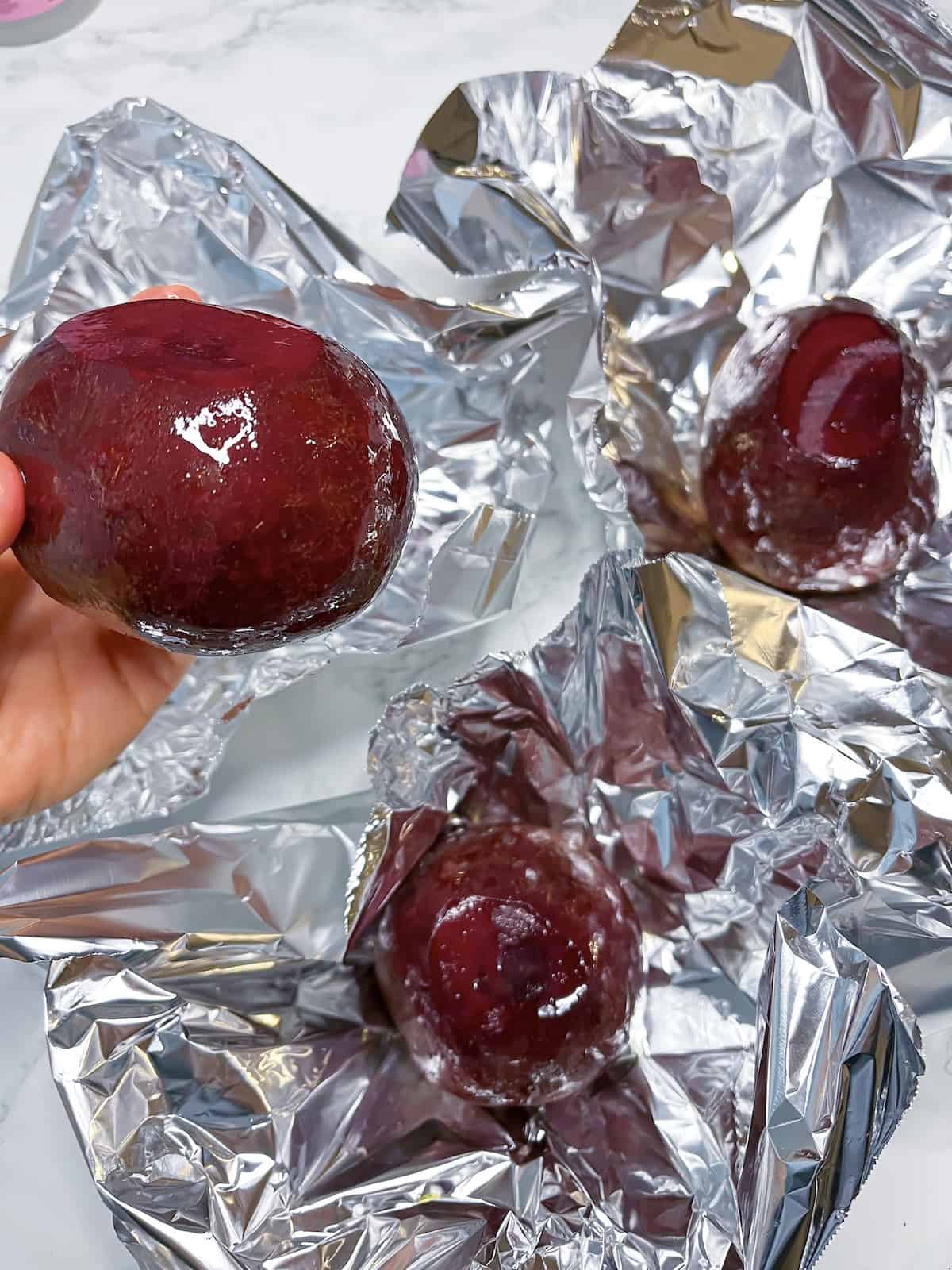 Raw beets coated in olive oil and being put in a foil packet to be roasted. A hand is holding one of them up
