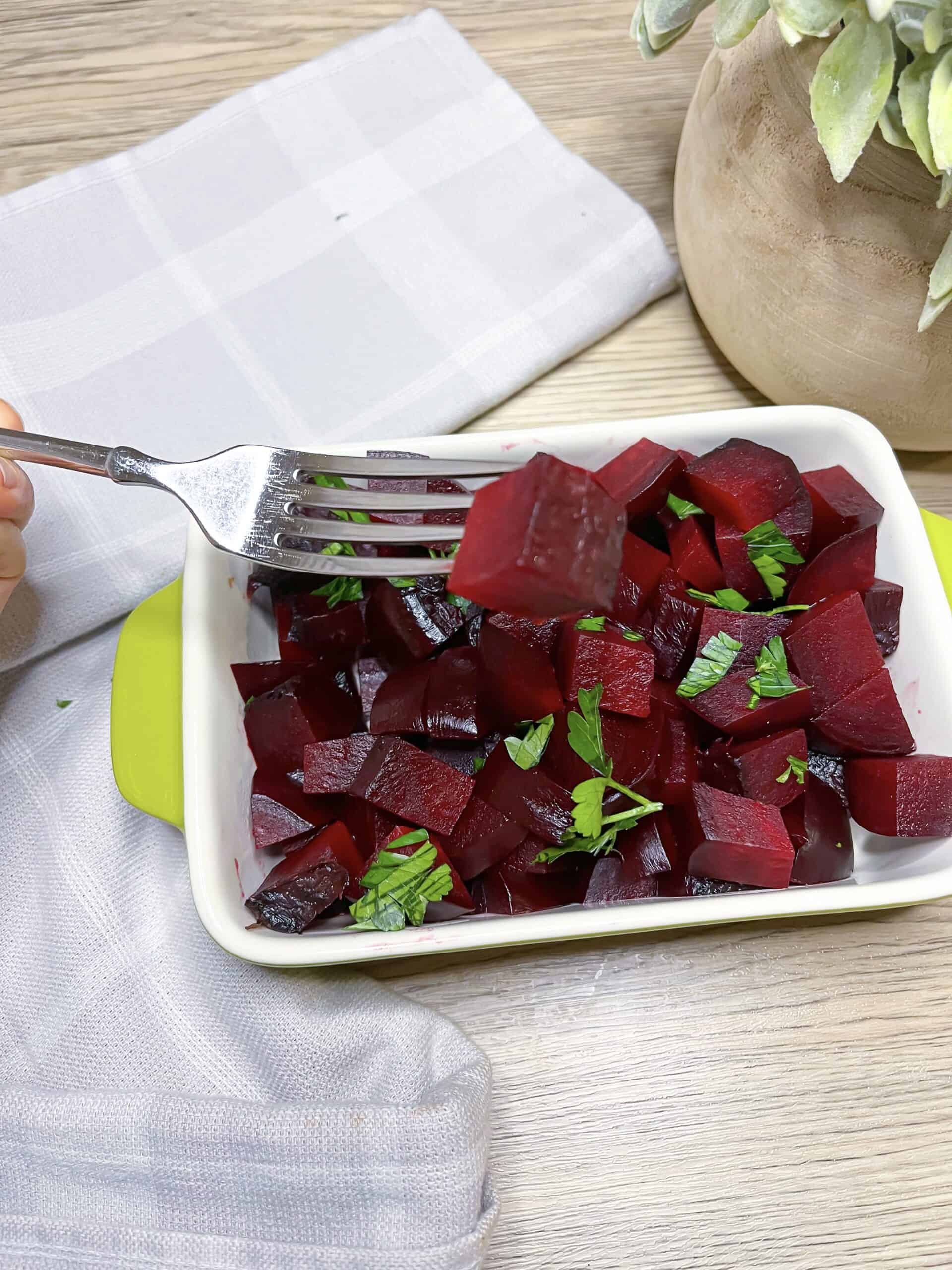 Roasted beets in foil diced with some parsley on top and a hand holding a fork that is piercing a beet with a gray dish cloth and green plant in the background