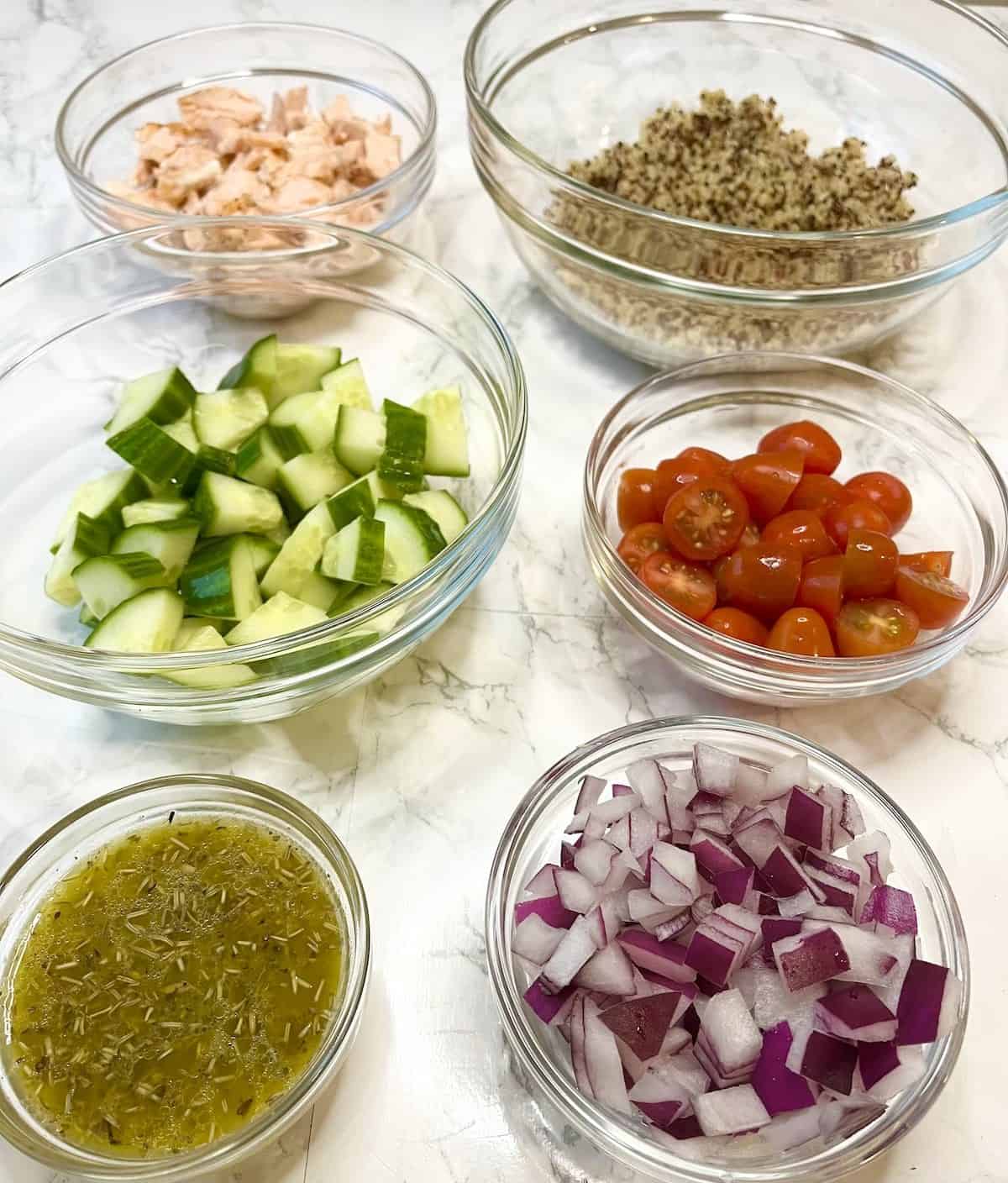 The ingredients on a white marble table including a bowl with salmon, quinoa, cucumbers, tomatoes, lemon herb dressing, and red onions