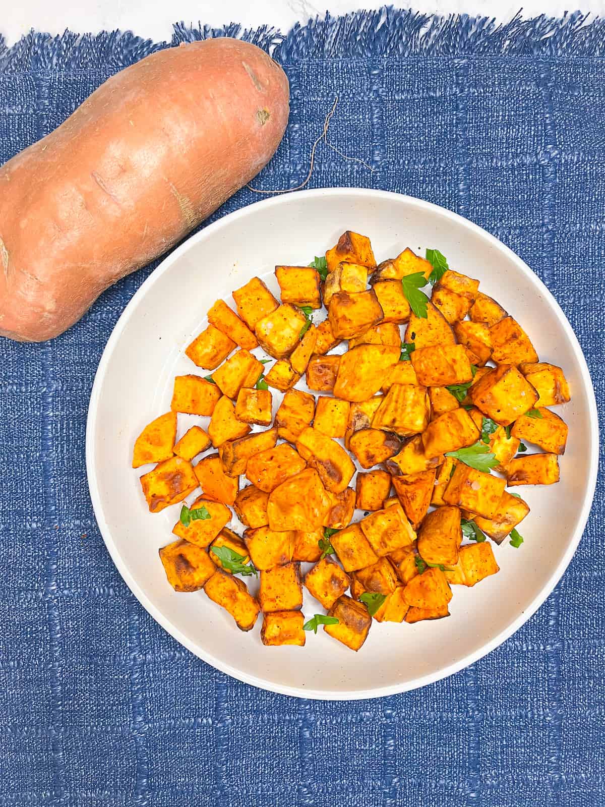 air fryer sweet potato cubes in a white bowl with a whole sweet potato in the background on a denim placemat