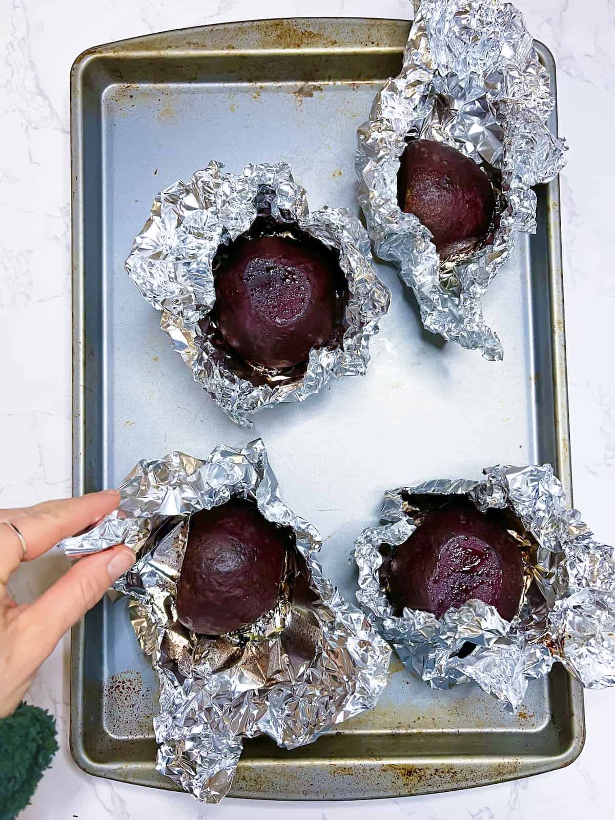 roasted beets in foil on a baking sheet with a hand holding one foil packet opened