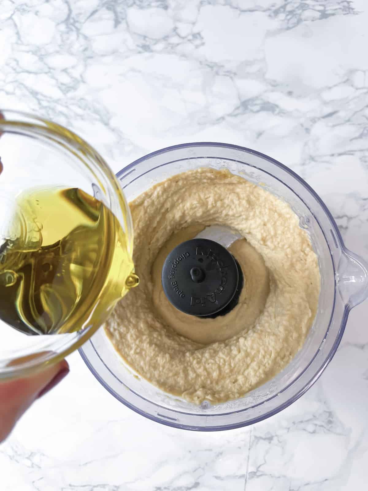 A hand pouring olive oil into a food processor with a chickpea and tahini and lemon juice mix