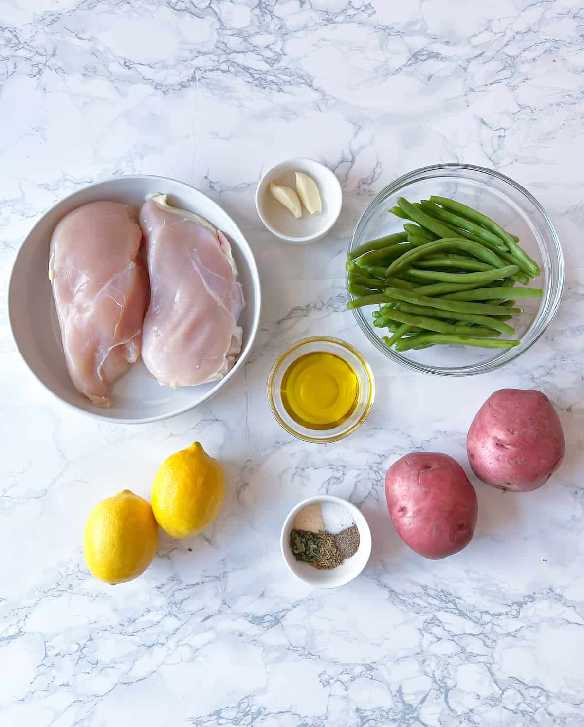 Ingredients for Slow Cooker Chicken, Green Beans and Potatoes on a marble table. There are large boneless skinless chicken breasts, fresh green beans, red potatoes, lemons, olive oil, and spices