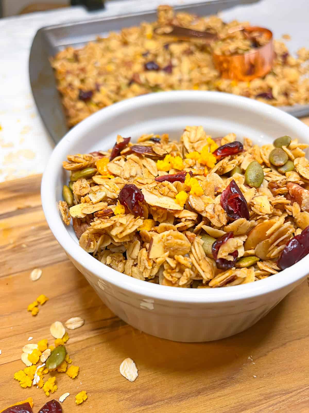 Orange granola with cranberries and pecans in a white bowl with more homemade granola on a baking sheet in the background