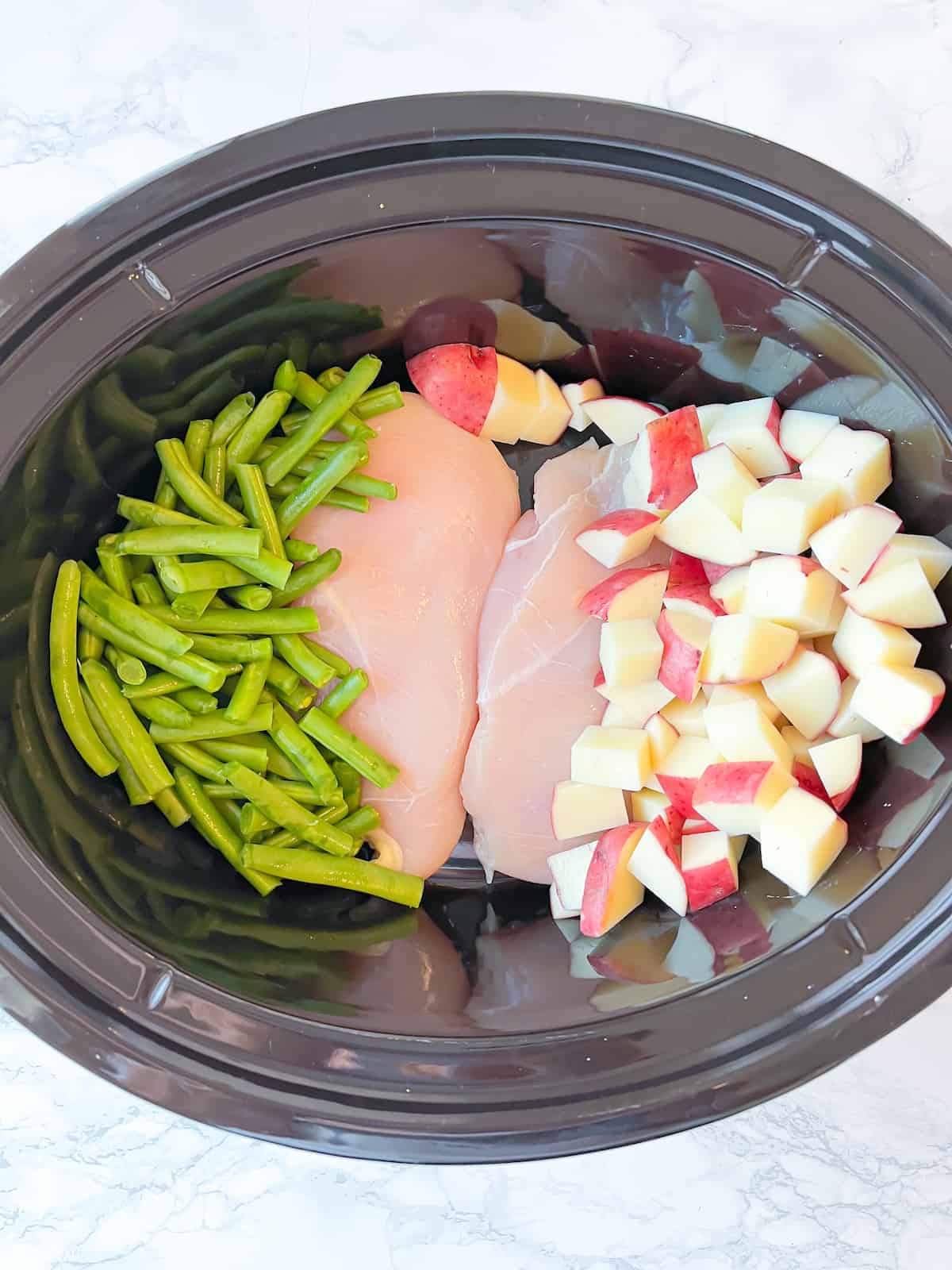 Slow cooker chicken and potatoes in the bottom of a crockpot with green beans and red potatoes stacked to the side of boneless, skinless chicken breasts which are in the center in an even layer