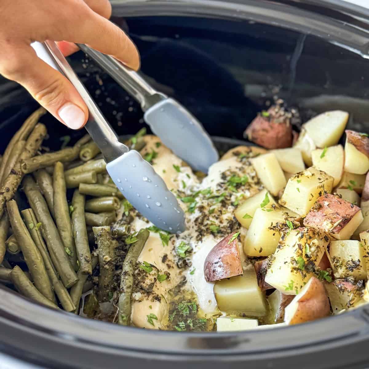 Human hand shredding chicken in a crockpot with potatoes and green beans.