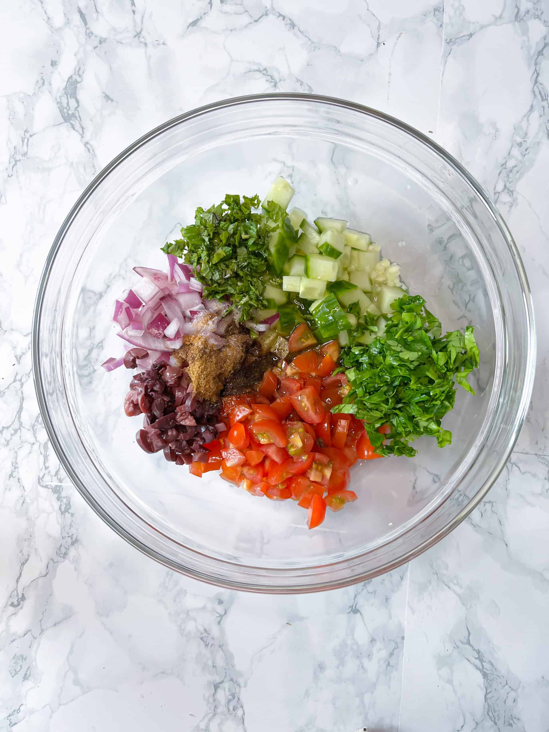 a glass mixing bowl with diced tomatoes, cucumber, red onion, kalamata olives, parsley and mint with spices and lemon juice and olive oil on top