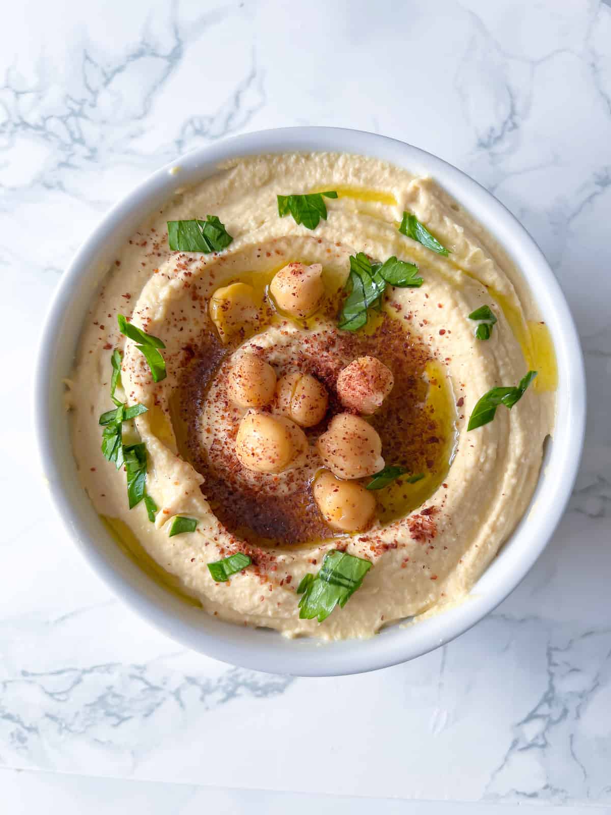 homemade hummus in a white bowl on a marble background with chickpeas and fresh parsley on top