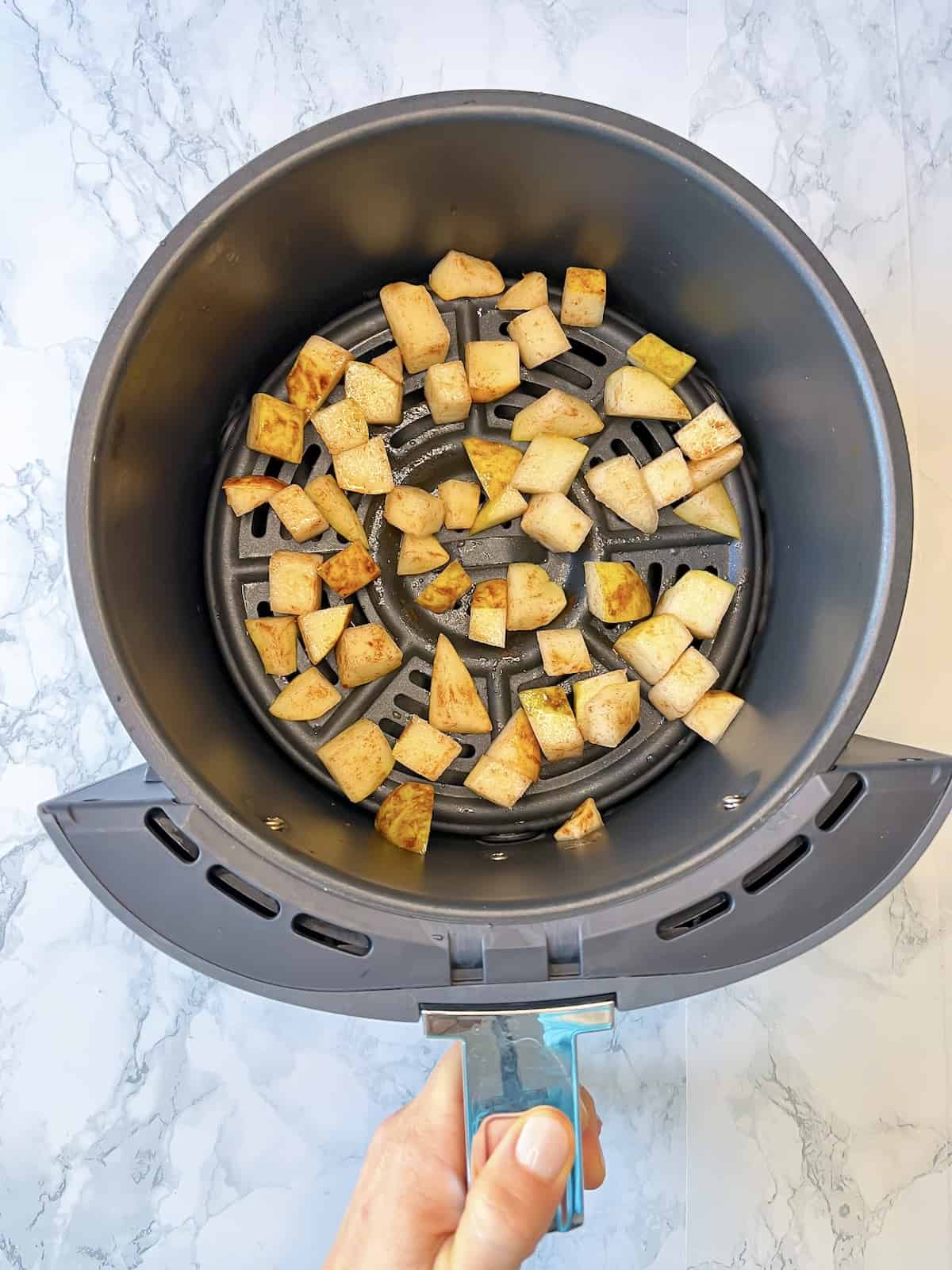 a hand holding the handle of an air fryer with diced pears in a single layer