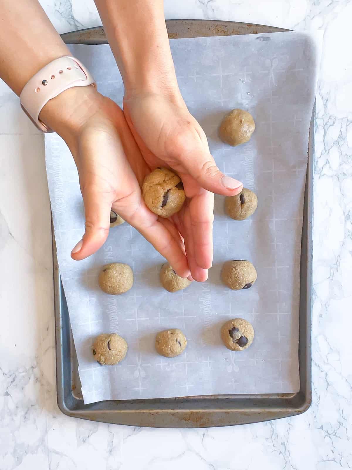 hands rolling a protein cookie dough ball with a baking sheet of them in the background