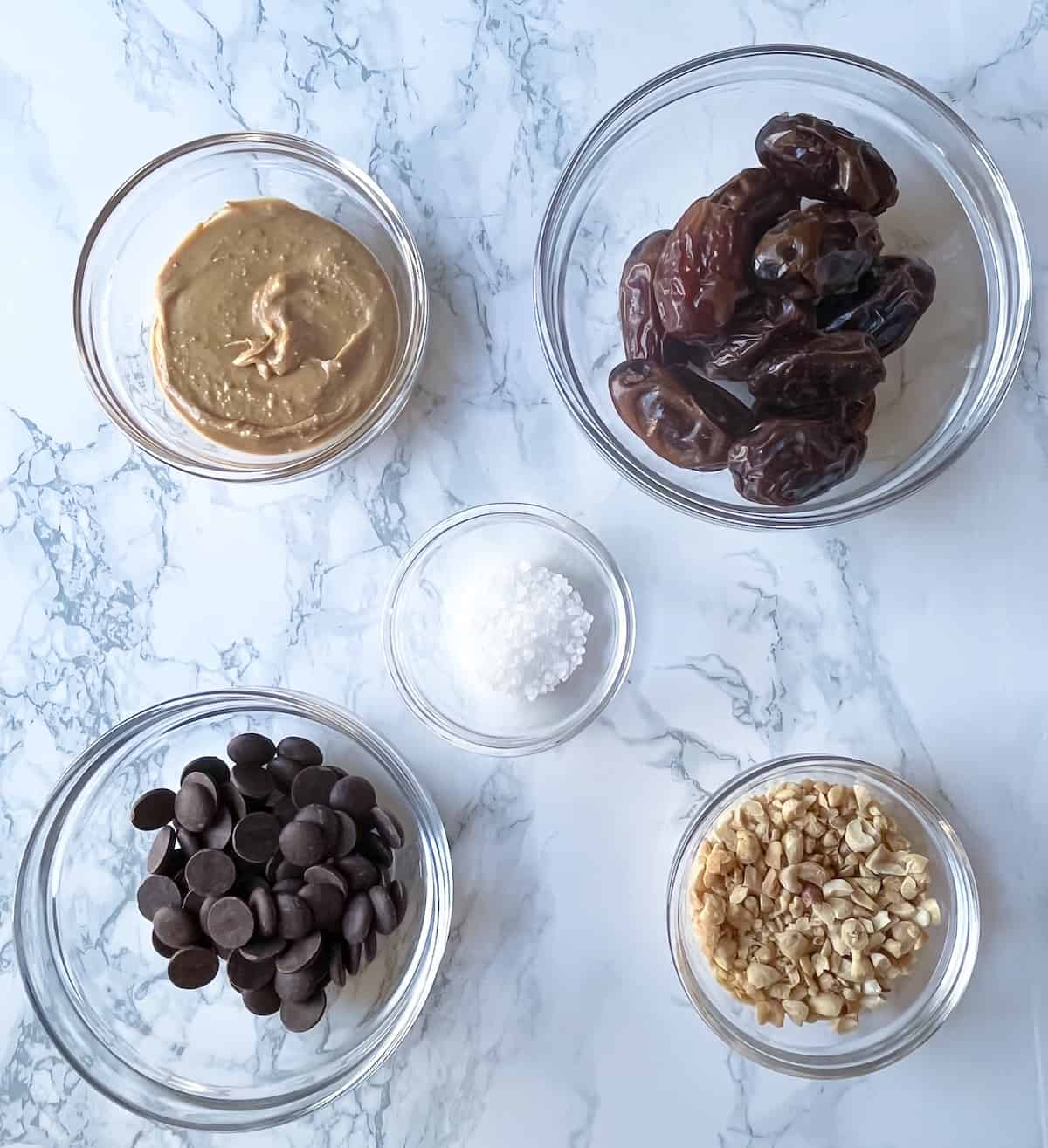 ingredients for date snickers on a marble counter including peanut butter, medjool dates, chocolate chips, flaky salt, and chopped peanuts