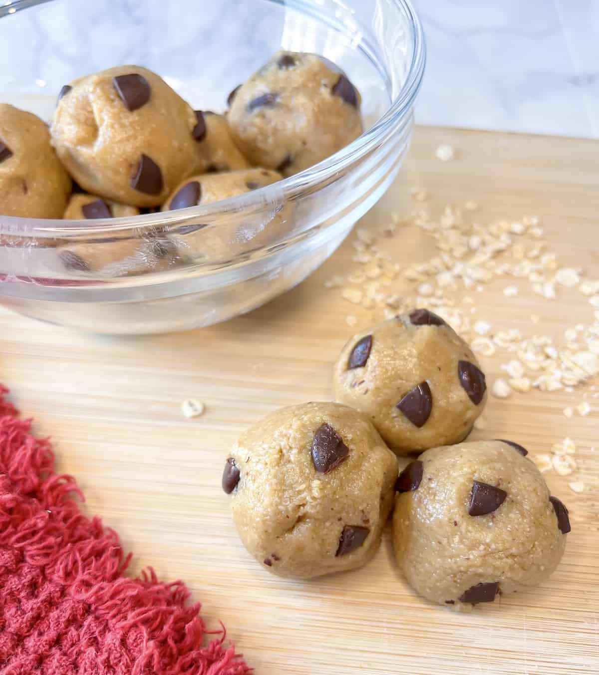 protein cookie dough bites on a wooden board with a glass bowl full of cookie dough in the background