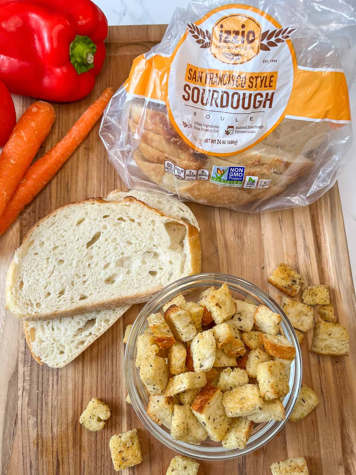 sourdough croutons in a small glass bowl with 2 slices of sourdough bread and a loaf of Izzio's sourdough bread in the background with a red bell pepper and 2 raw carrots