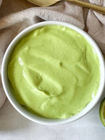 avocado lime crema in a white bowl with a lime next to it and a spoon in the background.