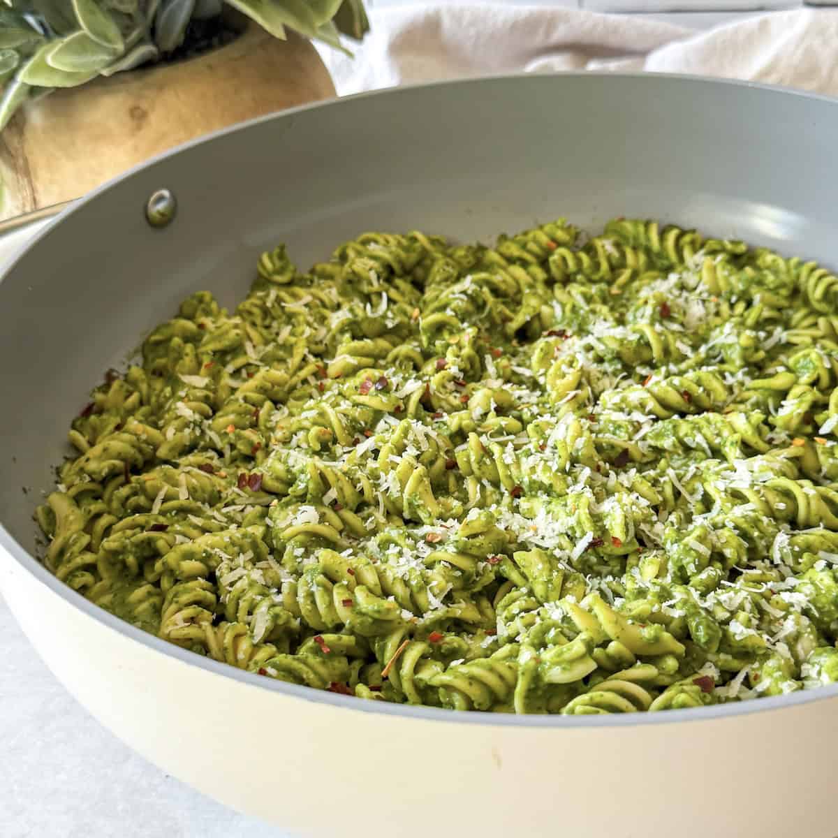 side view of green pasta sauce in pasta in a skillet with parmesan on top.
