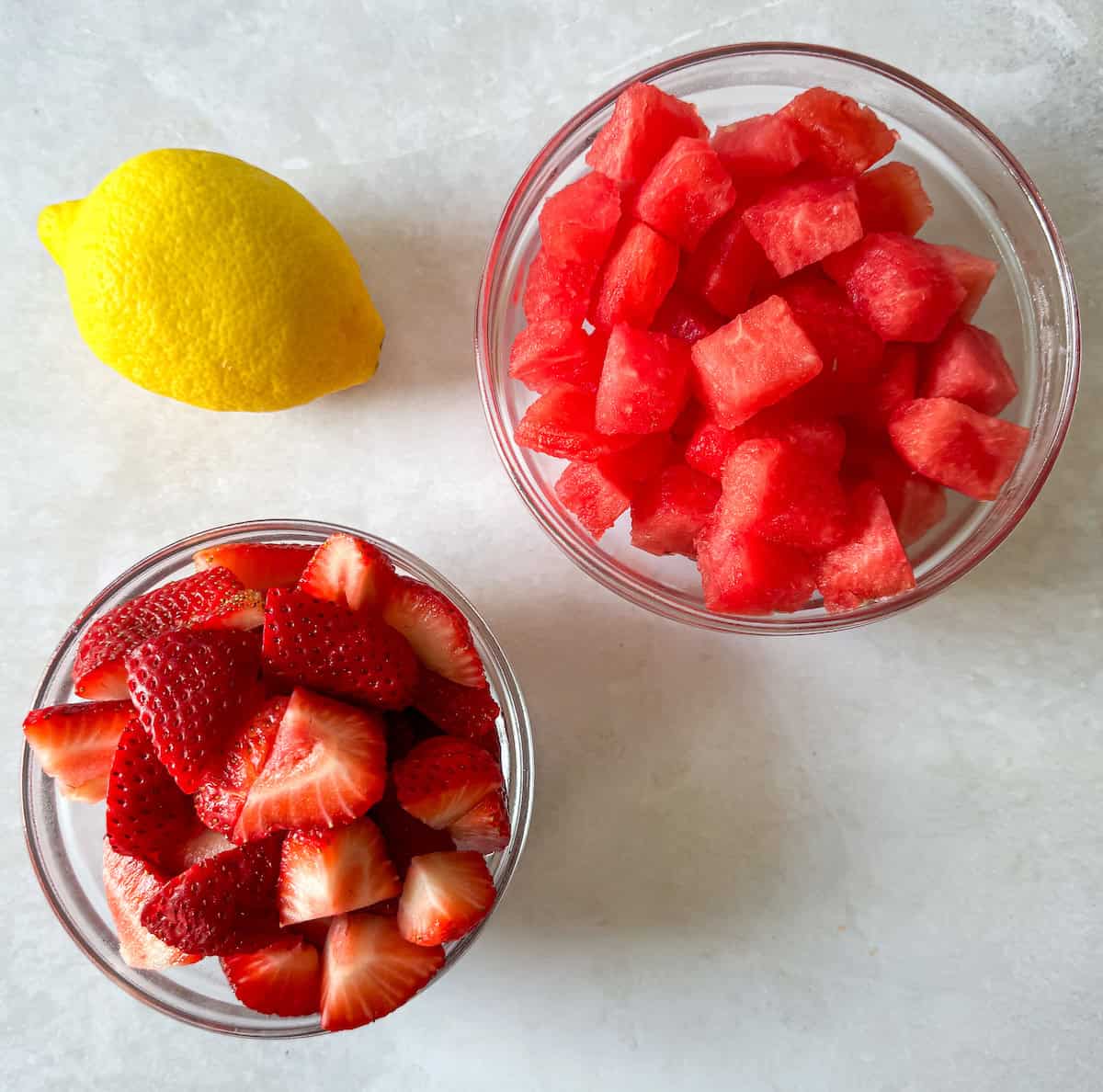 Ingredients to make this recipe on a table including fresh strawberries, watermelon cubes, and a lemon.