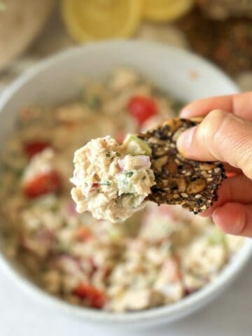 A human hand holding a seed cracker with mediterranean tuna salad on it.