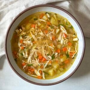 A bowl of chicken soup no noodles fresh after cooking in a big white soup bowl with a dark brown rim.