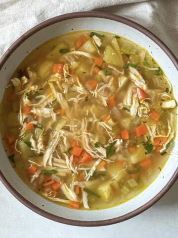 A bowl of chicken soup no noodles fresh after cooking in a big white soup bowl with a dark brown rim.
