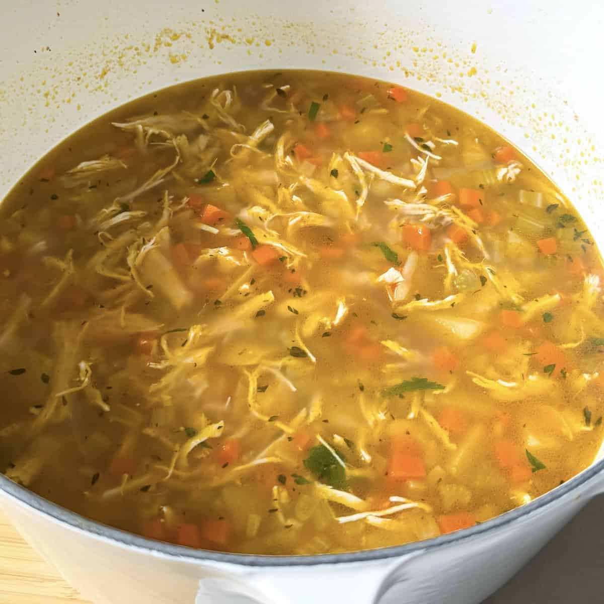 Shredded chicken added to a white dutch oven with cooked diced carrots, celery, and onion along with broth in a large white dutch oven.