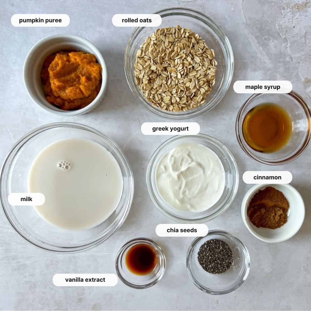 ingredients for pumpkin pie overnight oats in separate bowls on a gray and white marble background.
