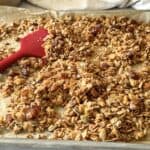 Apple pie granola on a baking sheet with a red spatula in the background.