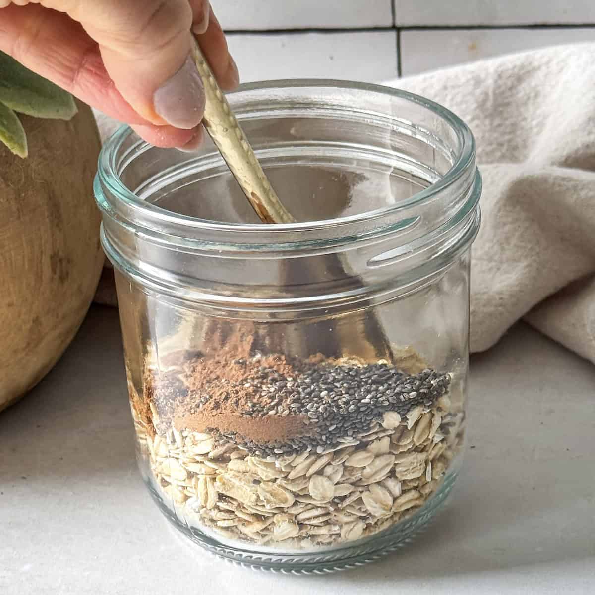 a human hand stirring oats, chia seeds, and cinnamon in a mason jar.