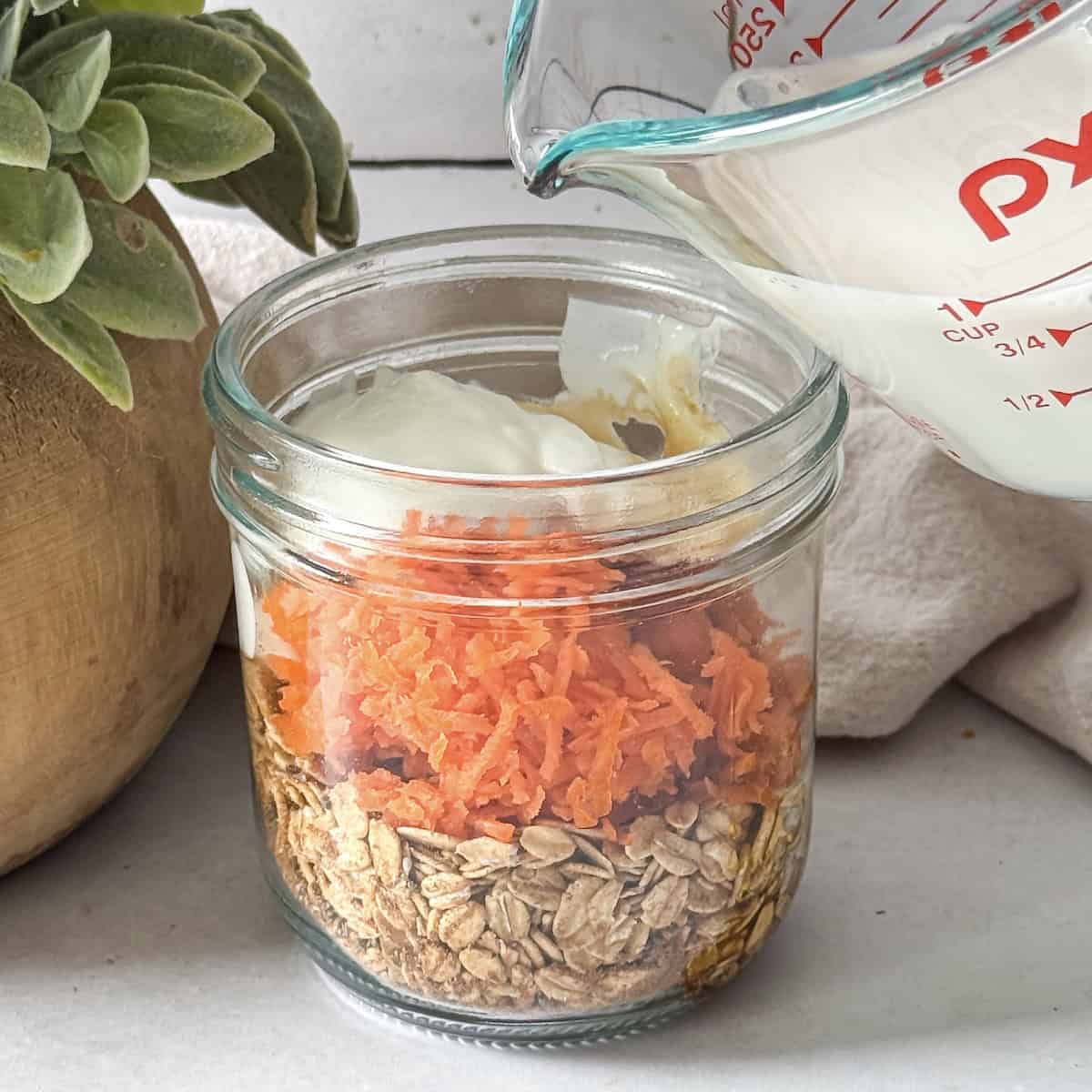 grated carrots and other wet ingredients in a mason jar with a human hand pouring milk into it.