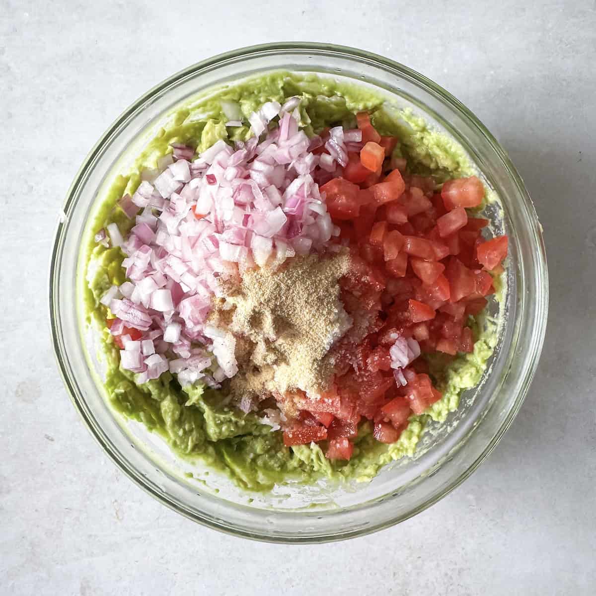 fresh ingredients for guacamole added to a glass bowl with mashed avocado (red onion, roma tomatoes, and garlic powder) overhead view.