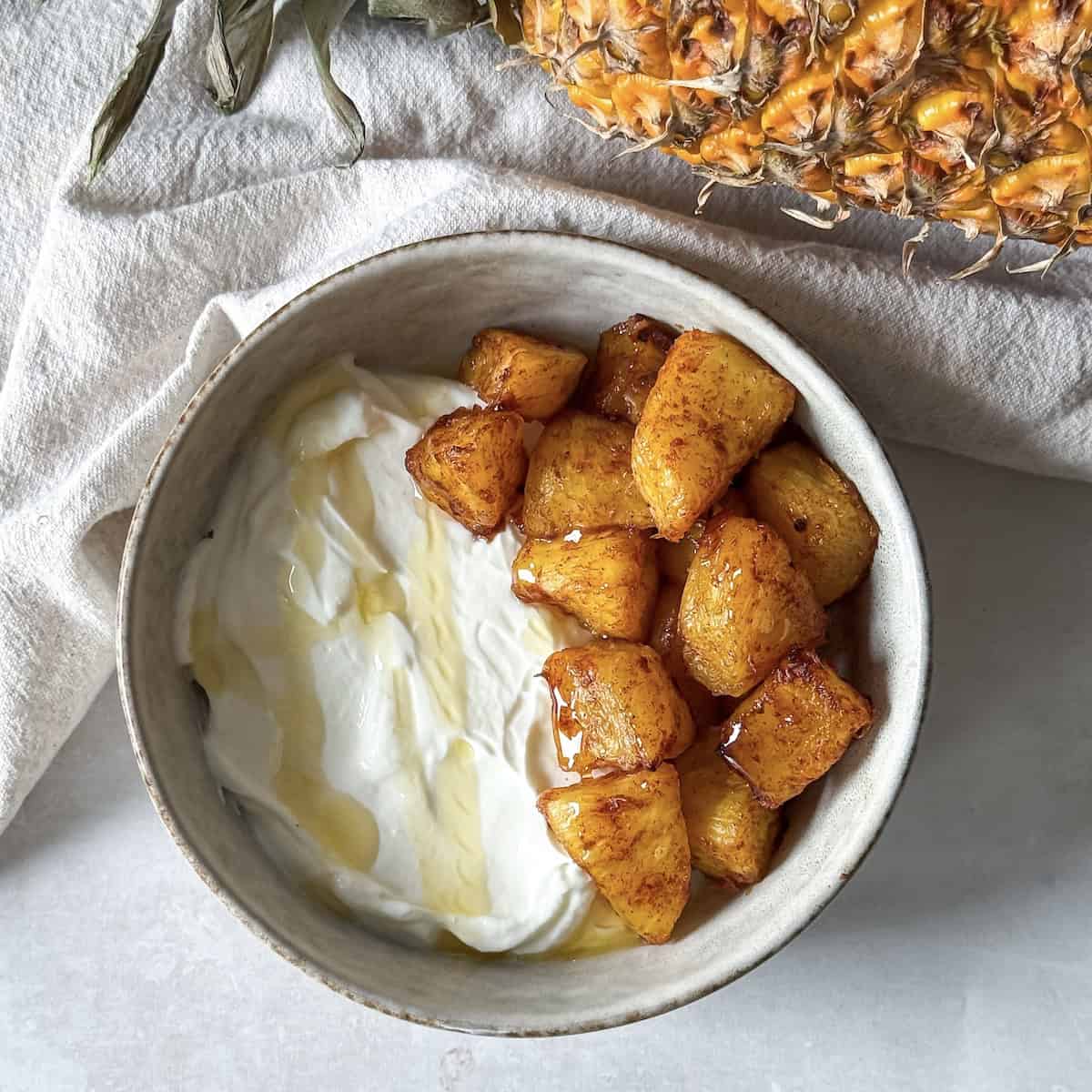 a bowl of air fryer pineapples in a bowl with Greek yogurt with a fresh pineapple in the background overhead view
