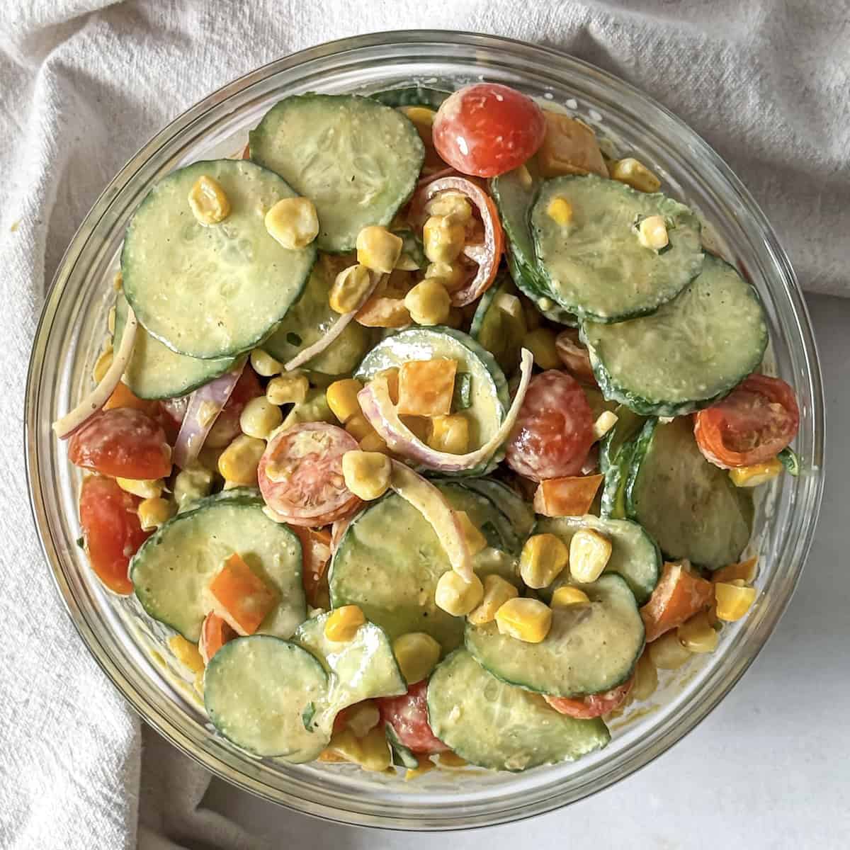 corn cucumber salad overhead view in a glass bowl with a dish cloth behind it.