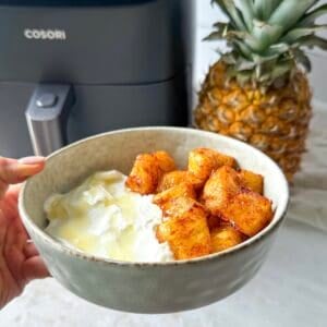 human hand holding a bowl of air fryer pineapples in a bowl with Greek yogurt with a fresh pineapple and air fryer in the background.