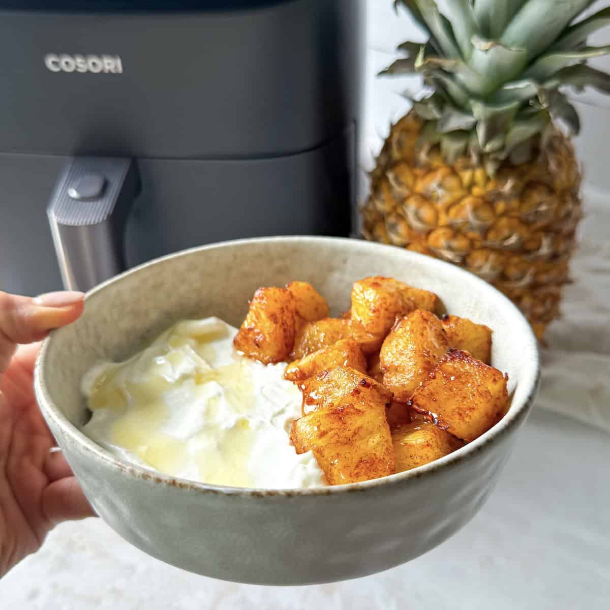 human hand holding a bowl of air fryer pineapples in a bowl with Greek yogurt with a fresh pineapple and air fryer in the background.
