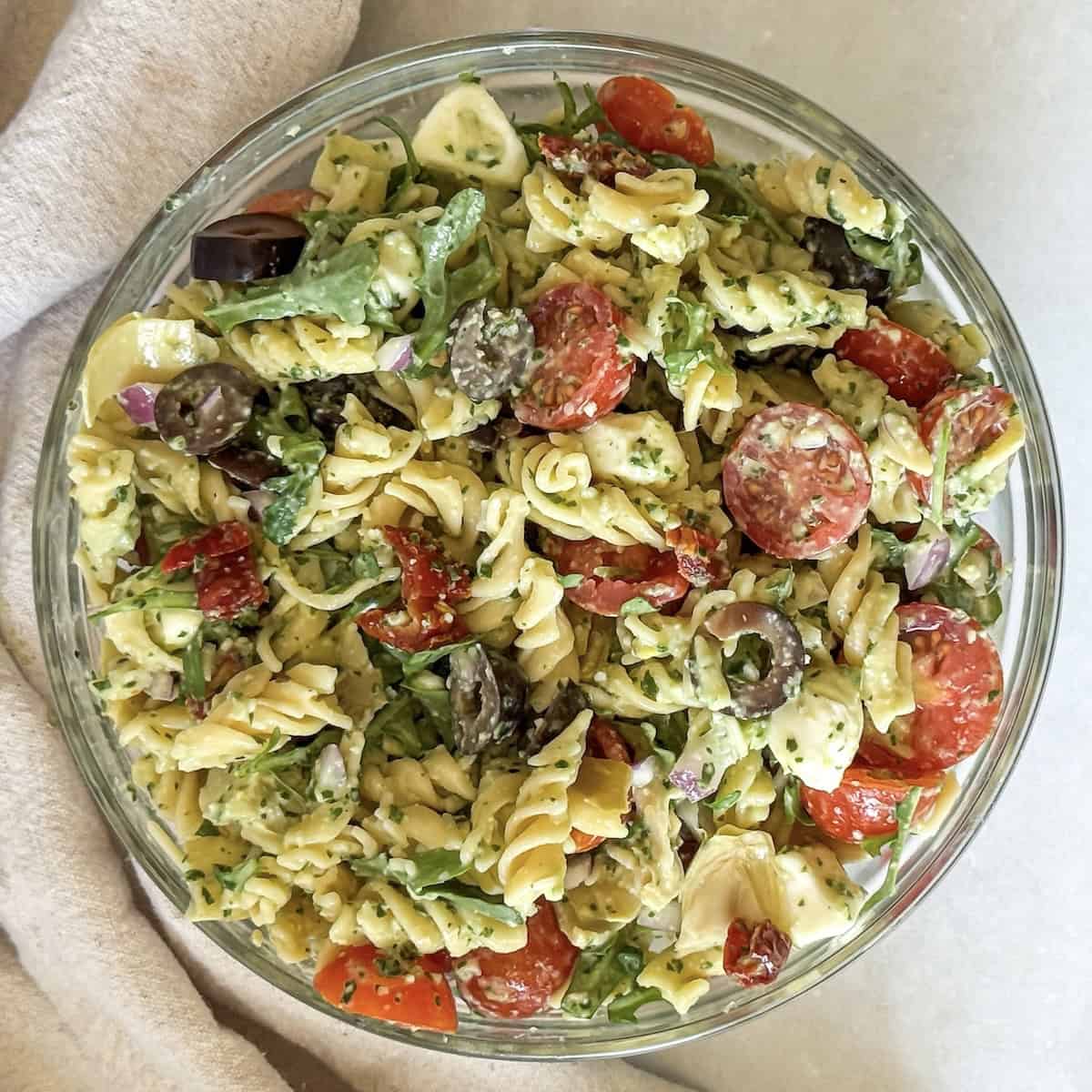 ingredients for pesto pasta salad with sundried tomatoes in a glass bowl overhead with a towel.