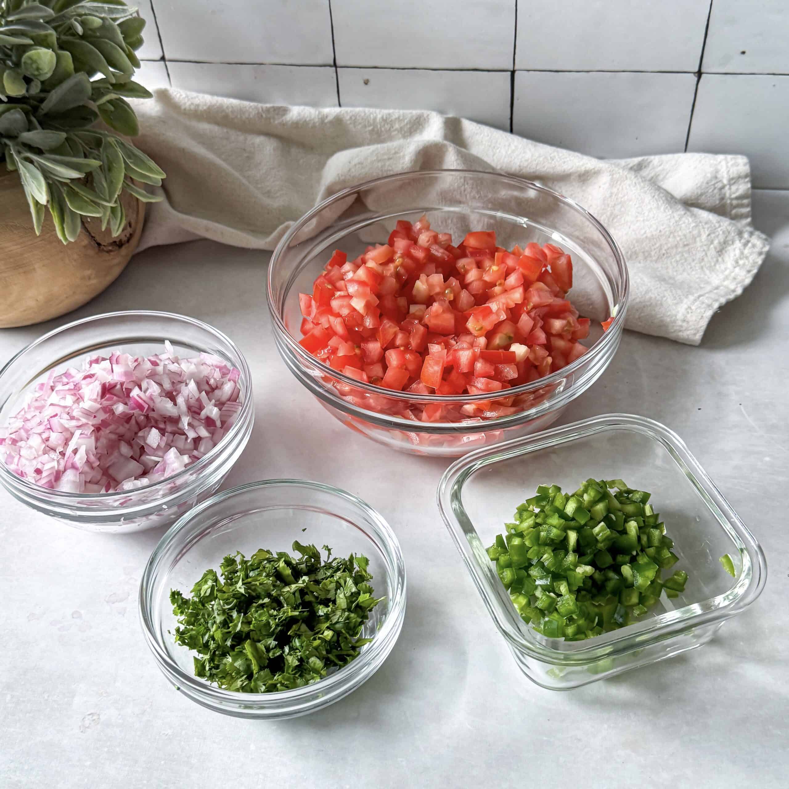 chopped tomtaotes, red onion, cilantro, and jalapeno in glass bowls.