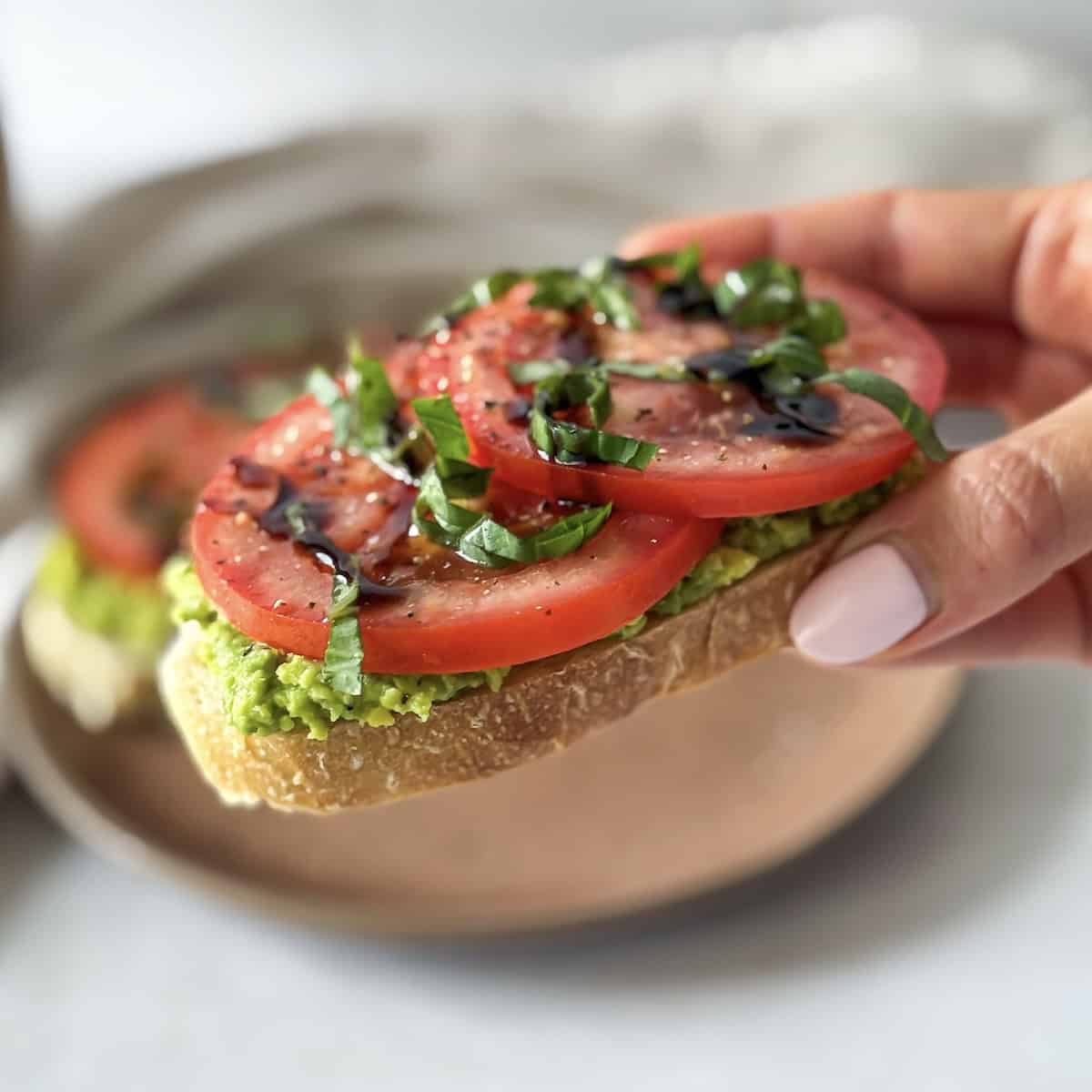 a human hand holding a slice of avocado tomato toast.