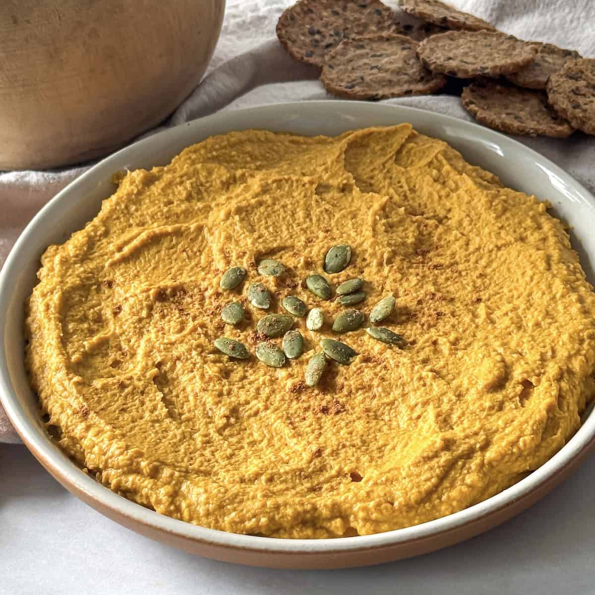 side view of pumpkin hummus on a plate with crackers in the background.