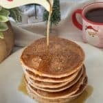 a human hand pouring maple syrup on top of a stack of gingerbread pancakes.