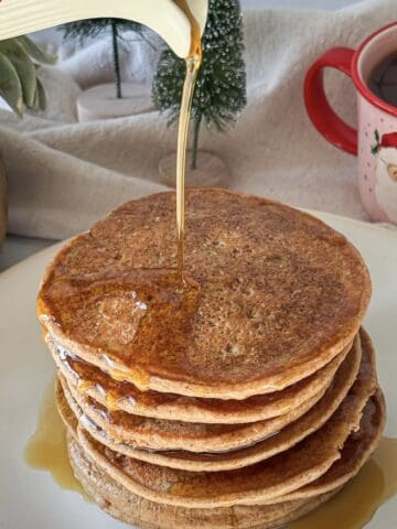 a human hand pouring maple syrup on top of a stack of gingerbread pancakes.