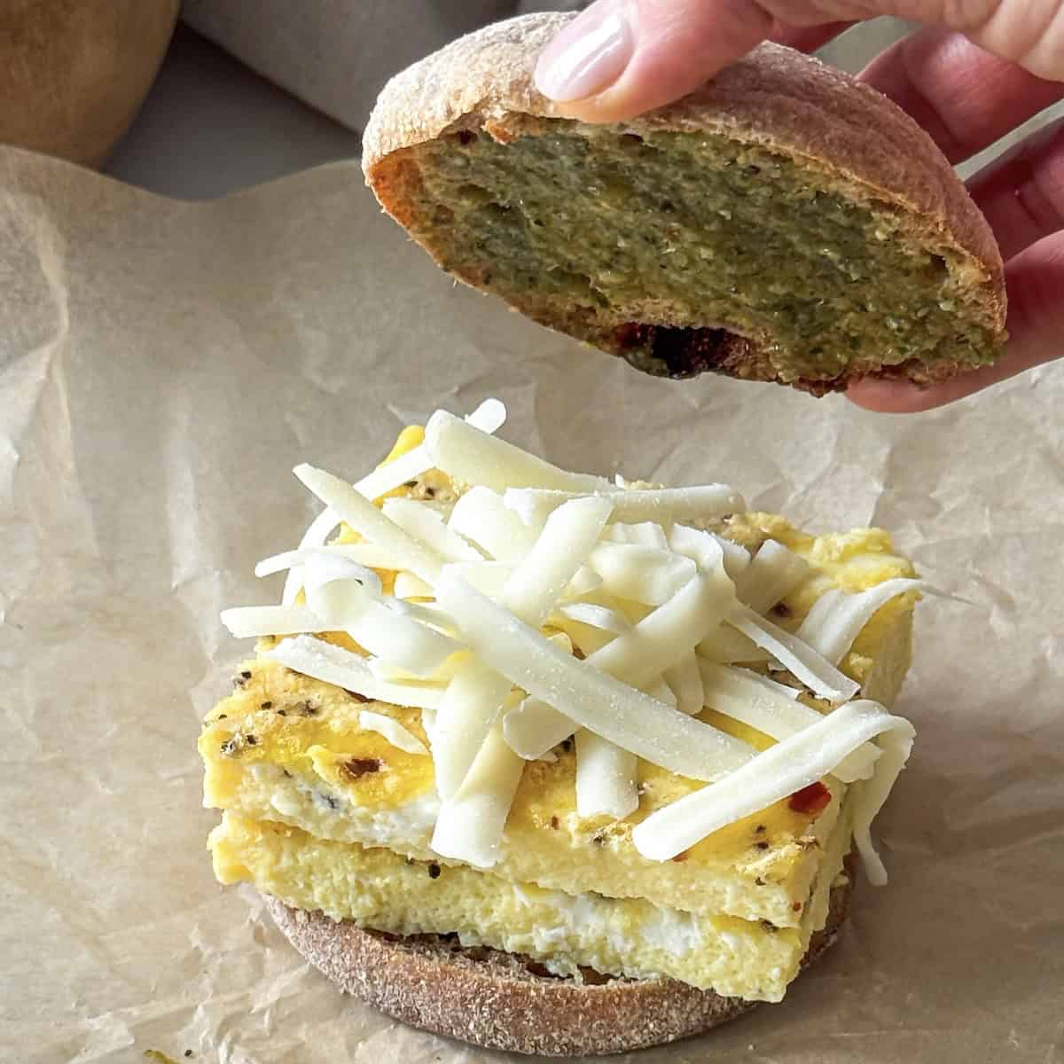 human hand putting an english muffin top with pesto on it on top of eggs.