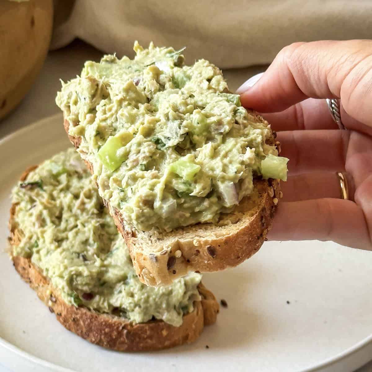 a human hand holding a slice of seeded toast with tuna salad with white beans on it.