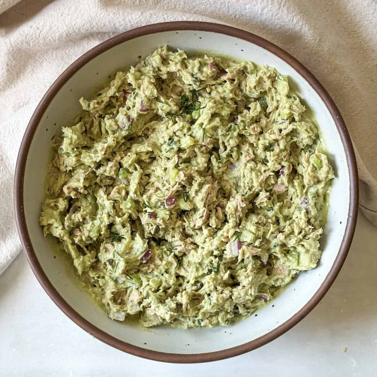 a white speckled bowl with a brown rim with a slightly vibrant green tuna salad after all the ingredients have been combined together.