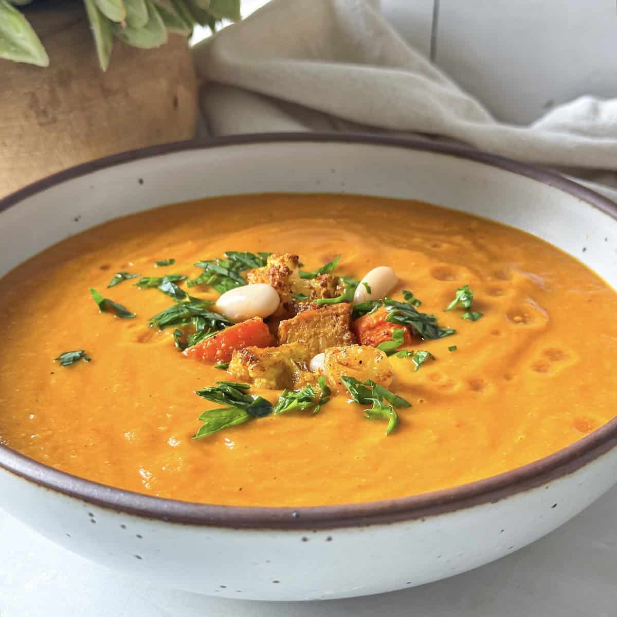 A golden soup in a big white bowl with brown speckles and a tan rim. The soup is garnished with chopped fresh parsley, white beans, and a couple pieces of roasted vegetables.